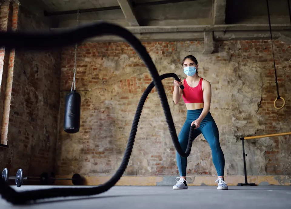 Young woman exercising with battle ropes at the gym wearing a medical mask.